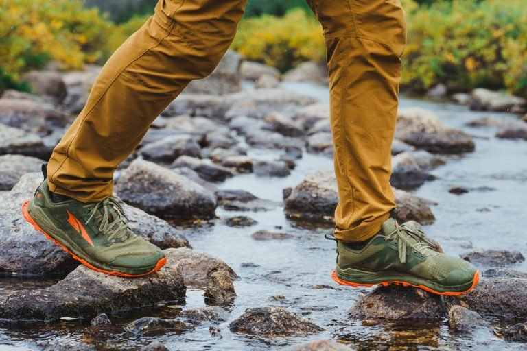 Multi-day hiker crossing a stream wearing trail running shoes and durable outdoor pants. The focus is on the footwear's grip and performance on wet, uneven rocks.